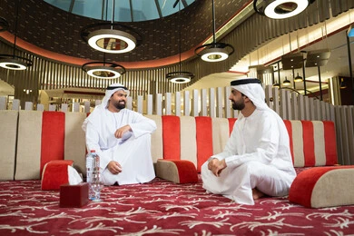 ايماءات كرم الضيافة وروح التواصل الاجتماعي في المجالس الإماراتية، تصميم داخلي أنيق بإضاءة دافئة ولمسات فخمة ،رجلان عربيان خليجيان إماراتيان يرتديان الكندورة والغترة، يجلسان في مجلس عصري بأجواء راقية وحديثة، يتبادلان الأحاديث الودية والابتسامات،مفهوم الاصالة والمعاصرة.