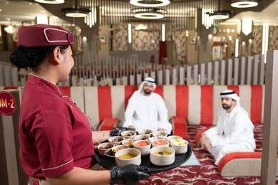 A waitress in a uniform is serving orders, generosity and hospitality among Arabs, spending enjoyable times with friends, two Arab Gulf men from the UAE wearing kandura and ghutrah sharing food.