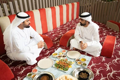 مفهوم الضيافة العربية والأجواء الاجتماعية، رجلان عربيان خليجيان إماراتيان يرتديان الكندورة والغترة يجلسان معًا داخل خيمه في أجواء دافئة ويتناولان وجبة تقليدية لذيذة من الأرز والمأكولات الشرقية،  مشاركة الطعام بروح الود والتقارب، خلفية داخلية مليئة بالتفاصيل الشرقية الجميلة.