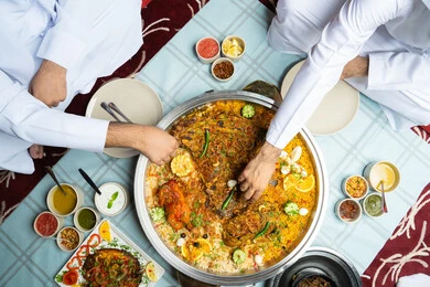 رجلان عربيان خليجيان إماراتيان يرتديان الكندورة والغترة يجلسان معًا داخل خيمه في أجواء دافئة ويتناولان وجبة تقليدية لذيذة من الأرز والمأكولات الشرقية، مفهوم الضيافة العربية والأجواء الاجتماعية، مشاركة الطعام بروح الود والتقارب، خلفية داخلية مليئة بالتفاصيل الشرقية الجميلة.