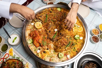 جلسة و كشتة بر شبابية، رجلان عربيان خليجيان إماراتيان يرتديان الكندورة والغترة يجلسان معًا داخل خيمه في أجواء دافئة ويتناولان وجبة تقليدية لذيذة من الأرز والمأكولات الشرقية، مفهوم الضيافة العربية والأجواء الاجتماعية، مشاركة الطعام بروح الود والتقارب، خلفية داخلية مليئة بالتفاصيل الشرقية الجميلة.