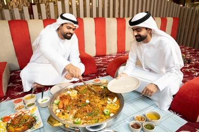 مشاركة الطعام بروح الود والتقارب، رجلان عربيان خليجيان إماراتيان يرتديان الكندورة والغترة يجلسان معًا داخل خيمه في أجواء دافئة ويتناولان وجبة تقليدية لذيذة من الأرز والمأكولات الشرقية، مفهوم الضيافة العربية والأجواء الاجتماعية، خلفية داخلية مليئة بالتفاصيل الشرقية الجميلة.