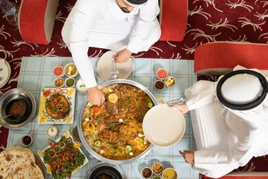 The concept of Arab hospitality and social atmosphere, two Emirati Gulf Arab men wearing kandura and ghutrah sitting together inside a tent in a warm environment, enjoying a delicious traditional meal of rice and Eastern dishes, sharing food in a spirit of friendliness and closeness, with an interior background filled with beautiful Eastern details.