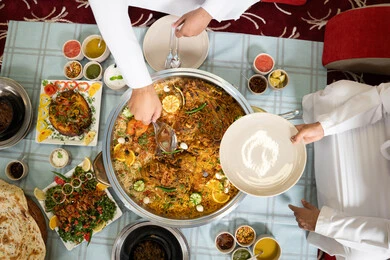 حسن الضيافة والاستقبال عند العرب ،رجلان عربيان خليجيان إماراتيان يرتديان الكندورة والغترة يجلسان معًا داخل خيمه في أجواء دافئة ويتناولان وجبة تقليدية لذيذة من الأرز والمأكولات الشرقية، مفهوم الضيافة العربية والأجواء الاجتماعية، مشاركة الطعام بروح الود والتقارب، خلفية داخلية مليئة بالتفاصيل الشرقية الجميلة.