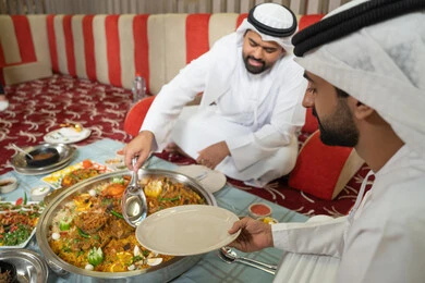A gathering and a youth outing, two Arab Gulf Emirati men wearing kandura and ghutrah sitting together inside a tent in a warm atmosphere, enjoying a delicious traditional meal of rice and Eastern dishes, embodying the concept of Arab hospitality and social ambiance, sharing food in a spirit of friendship and closeness, with an interior background filled with beautiful Eastern details.