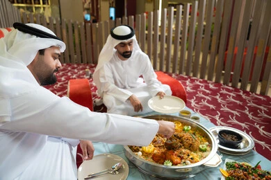 The concept of good hospitality and generosity, two Arab Gulf men from the UAE wearing kandura and ghutrah sitting together inside a tent in a warm atmosphere, enjoying a delicious traditional meal of rice and Eastern dishes, sharing food in a spirit of friendship and closeness, with an interior background filled with beautiful Eastern details.
