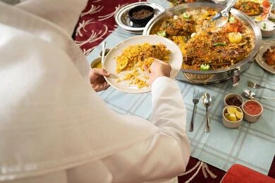 The concept of generosity and good hospitality, sharing food in a spirit of warmth and closeness, a close-up image of the hand of an Emirati Gulf Arab man wearing a kandura and ghutrah sitting at a table filled with delicious Eastern dishes, the values and traditions of Arab hospitality.