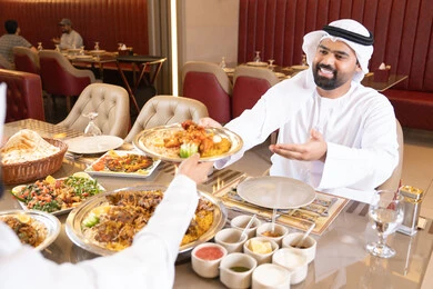 Two Arab Gulf Emirati men wearing kandura and ghutrah share food, enjoying lunch outdoors, spending pleasant times in the restaurant, restaurants and cafes of the UAE, gestures of happiness and joy.