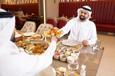 Restaurants and cafes in the UAE, enjoying lunch outdoors, spending enjoyable times in the restaurant, two Arab Gulf Emirati men wearing kandura and ghutrah sharing food, gestures of happiness and joy.