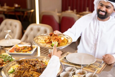 قضاء أوقات ممتعة في المطعم, الاستمتاع بالغداء في الخارج, مطاعم و كافيهات الامارات, رجلان عربيان خليجيان اماراتيان يرتديان الكندورة والغترة يتشاركان الطعام, إيماءات السعادة و السرور