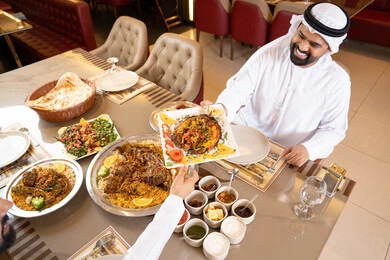 A fine restaurant in the United Arab Emirates, enjoying lunch outdoors, having a good time, restaurants and cafes in the UAE, two Arab Gulf Emirati men wearing kandura and ghutrah sharing food, gestures of happiness and joy.