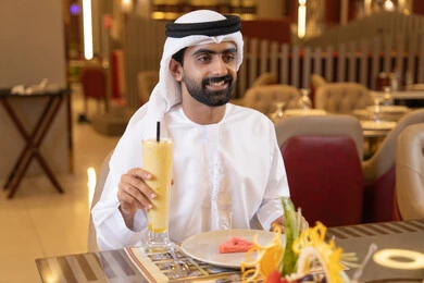 رجل عربي خليجي اماراتي يرتدي الكندورة والغترة يجلس على المائدة و يقوم بتناول الطعام, الاستمتاع بالغداء في الخارج, شرب المشروبات الباردة ،قضاء أوقات ممتعة في المطعم, مطاعم و كافيهات الامارات,إيماءات السعادة و السرور