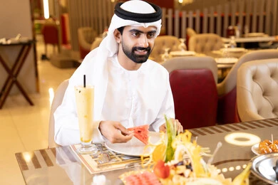 Spending enjoyable times at the restaurant, enjoying lunch outdoors, drinking cold beverages, restaurants and cafes in the UAE, an Arab Gulf Emirati man wearing the kandura and ghutrah sitting at the table and eating, gestures of happiness and joy.