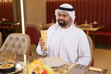 مطاعم و كافيهات الامارات, رجل عربي خليجي اماراتي يرتدي الكندورة والغترة يجلس على المائدة و يقوم بتناول الطعام, الاستمتاع بالغداء في الخارج, شرب المشروبات الباردة ،قضاء أوقات ممتعة في المطعم, إيماءات السعادة و السرور