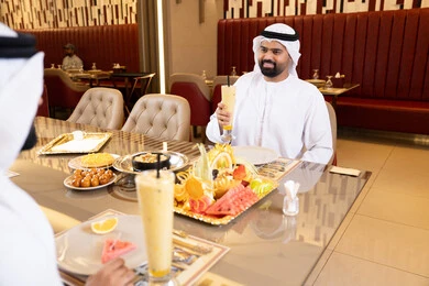 Spending enjoyable times at the restaurant, enjoying lunch outdoors, restaurants and cafes in the UAE, two Emirati Gulf Arab men wearing kandura and ghutrah sharing food, gestures of happiness and joy.