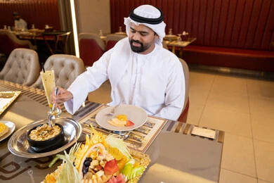 Drinking cold beverages, enjoying lunch outdoors, spending enjoyable times at the restaurant, restaurants and cafes in the UAE, an Arab Gulf Emirati man wearing the kandura and ghutrah sitting at the table and eating, gestures of happiness and joy.