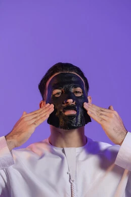 أهمية الفحم للبشرة, خطوات تنظيف وتقشير البشرة، الاستمتاع بلحظات العناية الذاتية، مفهوم التوازن المثالي بين النقاء والترطيب، بورتريه مقرب لرجل عربي خليجي اماراتي يرتدي الكندورة الاماراتية و يقوم بوضع ماسك على وجهه, خلفية بنفسجية