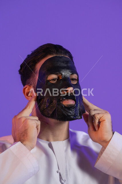 Steps for cleansing and exfoliating the skin, enjoying moments of self-care, the concept of the perfect balance between purity and hydration, a close-up portrait of an Emirati Gulf Arab man wearing an Emirati kandura and applying a mask to his face, purple background