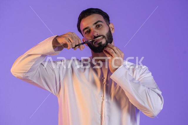 المحافظة على النظافة الشخصية، قص و تصفيف الذقن والشارب, مركز تجميلي رجالي، مفهوم العناية و الاهتمام بالجمال والمظهر الخارجي، بورتريه لرجل عربي خليجي اماراتي يرتدي الكندورة الاماراتية و يمسك المقص, خلفية بنفسجية