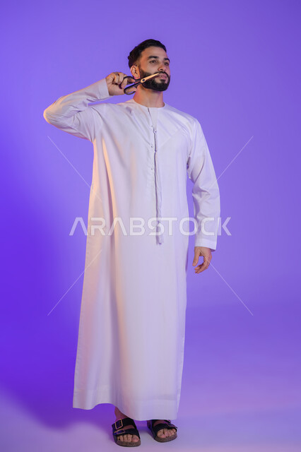 Beard and mustache trimming and styling, men's beauty center, concept of care and attention to beauty and appearance, portrait of an Emirati Gulf Arab man wearing the Emirati kandura and holding scissors, purple background