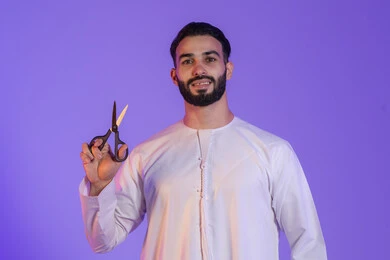 مركز تجميلي رجالي، مفهوم العناية و الاهتمام بالجمال والمظهر الخارجي، قص و تصفيف الذقن والشارب في صالون الحلاقة، بورتريه لرجل عربي خليجي اماراتي يرتدي الكندورة الاماراتية و يمسك المقص, خلفية بنفسجية