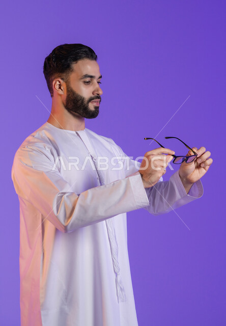 إيماءات التركيز والتمعن، مفهوم تحسين الرؤية ومحاولة تعديل البصر، الاهتمام بالمظهر الخارجي، ارتداء النظارات الخاصة بضعف النظر، بورتريه مقرب لرجل عربي خليجي اماراتي يرتدي الكندورة الاماراتية و ينظر للنظارة الطبية, خلفية بنفسجية