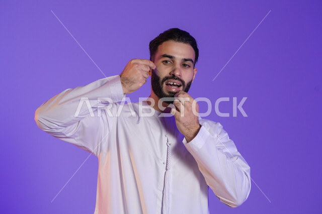 روتين صباحي للعناية بالفم, خطوة أساسية بعد كل وجبة للحفاظ على ابتسامة مشرقة, بورتريه لرجل عربي خليجي اماراتي يرتدي الكندورة الاماراتية و يقوم بتنظيف أسنانه باستخدام الخيط السني، مفهوم الاهتمام بصحة الأسنان، خلفية بنفسجية
