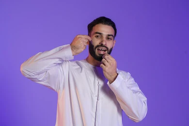 Morning oral care routine, an essential step after every meal to maintain a bright smile, portrait of an Emirati Gulf Arab man wearing an Emirati kandura and brushing his teeth using dental floss, concept of caring for dental health, purple background