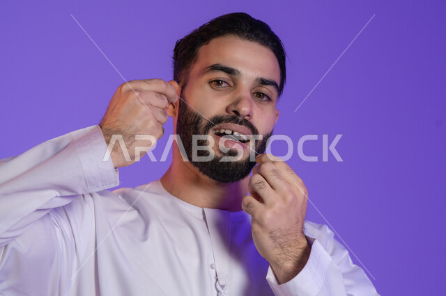 خطوة أساسية بعد كل وجبة للحفاظ على ابتسامة مشرقة, بورتريه لرجل عربي خليجي اماراتي يرتدي الكندورة الاماراتية و يقوم بتنظيف أسنانه باستخدام الخيط السني، مفهوم العناية بصحة الفم والأسنان، خلفية بنفسجية