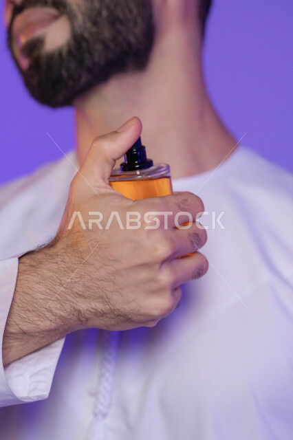 استخدام العود العطري، التطيب بالروائح الفريدة، استخدام الزيوت العطرية الطبيعية, مفهوم الاناقة والاهتمام بالمظهر الخارجي, التسويق لمنتجات العطور, بورتريه مقرب ليد شاب عربي خليجي اماراتي تمسك زجاجة عطر, خلفية بنفسجية