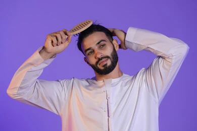 شعر صحي وأنيق، مفهوم الاهتمام بالجمال والأناقة والمظهر الخارجي،  بورتريه لرجل عربي خليجي اماراتي يرتدي الكندورة الاماراتية و يقوم بتصفيف شعره باستخدام الفرشاة, خلفية بنفسجية