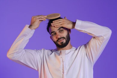 المحافظة على شعر صحي وأنيق، مفهوم الاهتمام بالجمال والأناقة والمظهر الخارجي، العناية بالنظافة الشخصية، بورتريه مقرب لرجل عربي خليجي اماراتي يرتدي الكندورة الاماراتية و يقوم بتصفيف شعره باستخدام الفرشاة, خلفية بنفسجية