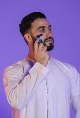 اختيار الأداة المناسبة للعناية باللحية والذقن, مفهوم الاعتناء بالبشرة والاهتمام بالمظهر الخارجي، المحافظة على الأناقة والنظافة، بورتريه لرجل عربي خليجي اماراتي يرتدي الكندورة الاماراتية و يمسك شفرة حلاقة, خلفية بنفسجية