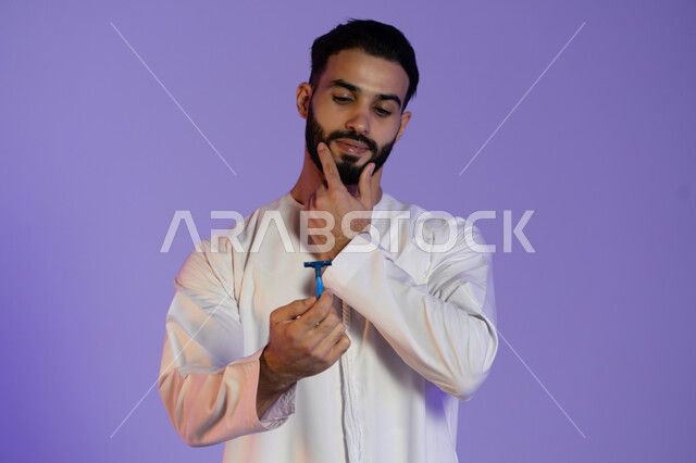 لحظة تركيز قبل الحلاقة, الحفاظ على الأناقة والنظافة الشخصية، العناية بالبشرة والاهتمام بالمظهر الخارجي، بورتريه لرجل عربي خليجي اماراتي يرتدي الكندورة الاماراتية و يمسك الشفرة, خلفية بنفسجية
