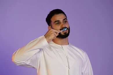 الانتعاش والوقاية الصحية, الاهتمام بالصحة والمظهر اليومي, الحفاظ على الأناقة والنظافة الشخصية، المحافظة على ابتسامة بيضاء، بورتريه لرجل عربي خليجي اماراتي يرتدي الكندورة الاماراتية و ينظف أسنانه بالفرشاة، خلفية بنفسجية