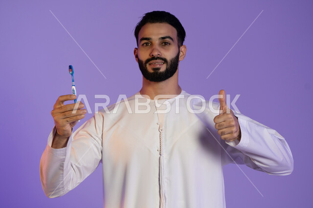 الشعور بالقبول والرضا، علامة التميز و الجودة, الحفاظ على الأناقة والنظافة الشخصية، العناية بالبشرة والاهتمام بالمظهر الخارجي، بورتريه لرجل عربي خليجي اماراتي يرتدي الكندورة الاماراتية يمسك الفرشاة يرفع اصبع الابهام باشارة اللايك، خلفية بنفسجية