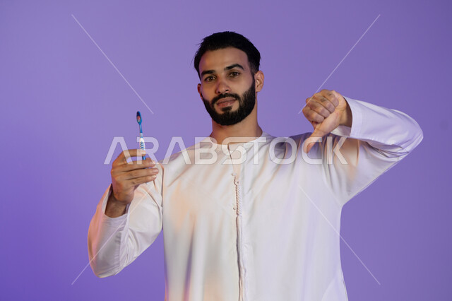 الشعور بالإنزعاج والغضب، حماية السن من التسوس، مفهوم العناية بصحة ونظافة الفم، بورتريه لرجل عربي خليجي اماراتي يرتدي الكندورة الاماراتية يمسك فرشاة الأسنان و يشير بأصبع الإبهام لأسفل, خلفية بنفسجية