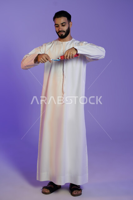 Protecting teeth from decay, the concept of oral health and hygiene, interest in elegance and appearance, maintaining a white smile, portrait of an Emirati Gulf Arab man wearing the Emirati kandura and holding a toothbrush and toothpaste, purple background