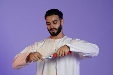 الإهتمام بصحة الأسنان، مفهوم العناية بصحة ونظافة الفم، الاعتناءبالأناقة والمظهر الخارجي، المحافظة على ابتسامة بيضاء، بورتريه لرجل عربي خليجي اماراتي يرتدي الكندورة الاماراتية و يمسك الفرشاة والمعجون، خلفية بنفسجية