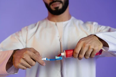 تجميل وتبييض الأسنان، مفهوم العناية بصحة ونظافة الفم، الاهتمام بالأناقة والمظهر الخارجي، المحافظة على ابتسامة بيضاء، بورتريه مقرب ليد رجل عربي خليجي اماراتي يرتدي الكندورة الاماراتية و يمسك  الفرشاة و المعجون، خلفية بنفسجية