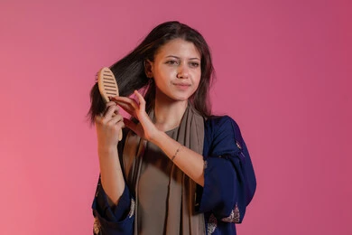 الاستعداد والتجهز للذهاب الى مكان ما، تصفيف الشعر والاعتناء بمظهره, الاهتمام بالجمال والأناقة، العناية بالنظافة الشخصية، بورتريه لامرأة عربية خليجية اماراتية مبتسمة ترتدي عباءة زرقاء و تقوم بتمشيط شعرها باستخدام الفرشاة، خلفية وردية