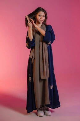 The concept of femininity and softness, hair styling and grooming, interest in beauty and elegance, personal hygiene, portrait of a smiling Emirati Gulf Arab woman wearing a blue abaya and brushing her hair, pink background