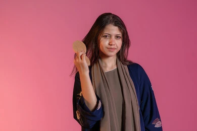 مفهوم التزين والاطلالة المتألقة، تصفيف الشعر والاعتناء بمظهره, الاهتمام بالجمال والأناقة، العناية بالنظافة الشخصية، بورتريه لامرأة عربية خليجية اماراتية مبتسمة ترتدي عباءة زرقاء و تقوم بتمشيط شعرها باستخدام الفرشاة، خلفية وردية