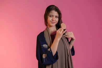 نظرات الأنوثة والنعومة، تصفيف الشعر والعناية بمظهره, مفهوم الاهتمام بالجمال والأناقة والمظهر الخارجي، العناية بالنظافة الشخصية، بورتريه لامرأة عربية خليجية اماراتية مبتسمة ترتدي عباءة زرقاء و تقوم بتمشيط شعرها باستخدام الفرشاة، خلفية وردية