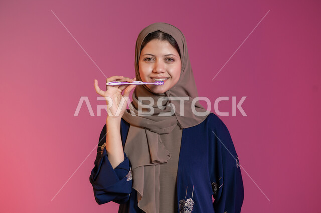 ابتسامة ناصعة البياض, روتين نظافة الفم اليومية، تجنب التسوس وأمراض اللثة, التأكد من النظافة بعد تناول الطعام, بورتريه لامرأة عربية خليجية اماراتية محجبة ترتدي عباءة زرقاء تمسك بيدها فرشاة الأسنان, خلفية وردية