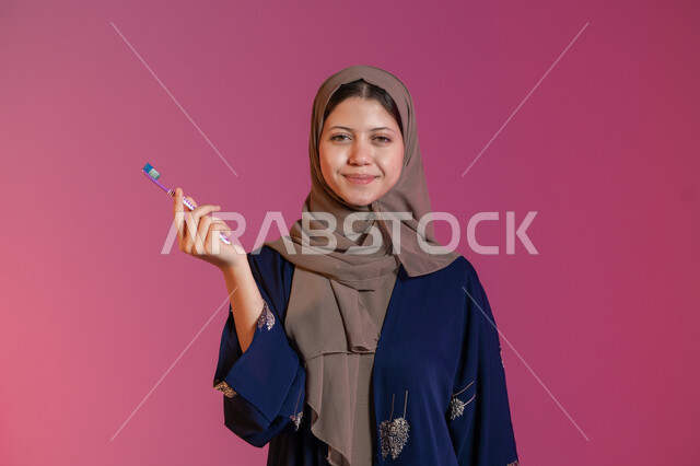 النظر إلى الكاميرا بإيماءات السرور، روتين نظافة الفم اليومية، تجنب التسوس وأمراض اللثة, التأكد من النظافة بعد تناول الطعام, بورتريه لامرأة عربية خليجية اماراتية محجبة ترتدي عباءة زرقاء تمسك بيدها فرشاة الأسنان, خلفية وردية