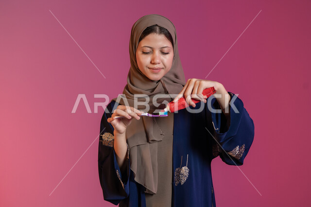 استخدام الفرشاة والمعجون، مفهوم العناية بصحة ونظافة الفم، الاهتمام بالأناقة والمظهر الخارجي، المحافظة على ابتسامة بيضاء، بورتريه لامرأة عربية خليجية اماراتية محجبة ترتدي عباءة زرقاء تقوم بتنظيف أسنانها, خلفية وردية