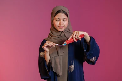 استخدام الفرشاة والمعجون، مفهوم العناية بصحة ونظافة الفم، الاهتمام بالأناقة والمظهر الخارجي، المحافظة على ابتسامة بيضاء، بورتريه لامرأة عربية خليجية اماراتية محجبة ترتدي عباءة زرقاء تقوم بتنظيف أسنانها, خلفية وردية