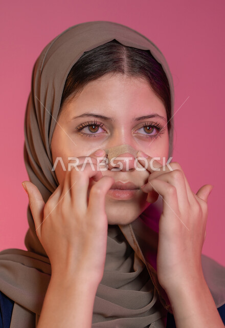 الاستمتاع بلحظات العناية الذاتية، استخدام مكونات تجميلية،  مفهوم العناية والاهتمام بنضارة وتحسين البشرة، التخلص من الرؤوس السوداء والجلد الميت، بورتريه مقرب لامرأة عربية خليجية اماراتية محجبة تقوم بوضع لصقات الأنف، خلفية وردية