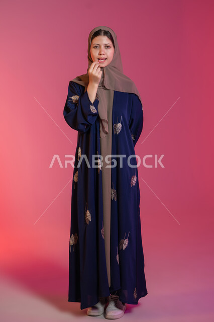 The concept of oral health and hygiene, interest in elegance and appearance, maintaining a white smile, portrait of a veiled Emirati Gulf Arab woman wearing a blue abaya, cleaning her teeth using dental floss, pink background